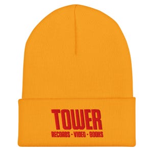May include: Mustard yellow beanie with a folded brim. The front of the beanie features the red text "TOWER" above "RECORDS VIDEO BOOKS". The beanie is made of a soft, knitted material and is a warm accessory.
