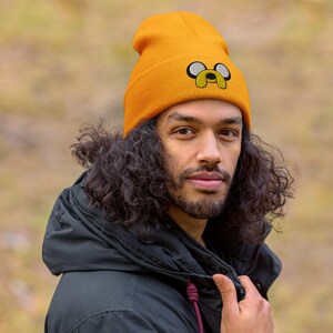 May include: A person wearing a yellow beanie with a cartoon dog face embroidered on the front. The beanie is made of a soft, knitted fabric and has a fold-over cuff.