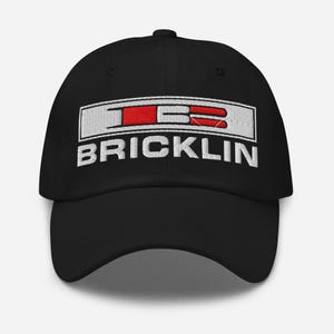 May include: Black baseball cap with the word "BRICKLIN" in white block letters. Above the word is a white rectangle with the letters "TB" in red and white. The cap has a curved brim and a button on top.