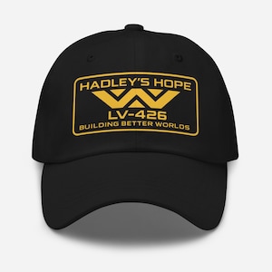 Op de afbeelding: Zwarte baseballcap met een gouden rechthoekige patch. De patch toont de tekst "HADLEY'S HOPE" boven een gestileerde "W" en de tekst "LV-426 BUILDING BETTER WORLDS". De cap heeft een gebogen klep en een verstelbare band.