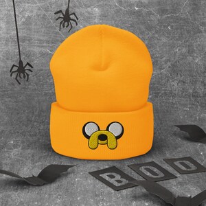 May include: A bright orange beanie with a black embroidered cartoon dog face. The beanie has a fold-over cuff.