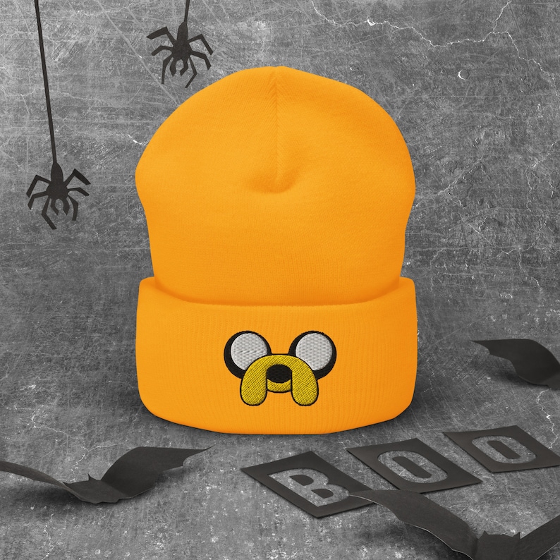 May include: A bright orange beanie with a black embroidered cartoon dog face. The beanie has a fold-over cuff.
