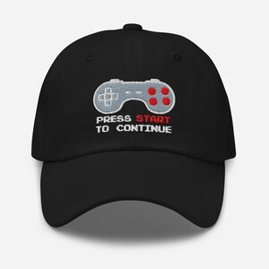 Puede incluir: Gorra de béisbol negra con un mando de videojuego gris bordado, botones rojos y el texto "PRESS START TO CONTINUE". La gorra tiene una visera curva y un ajuste estructurado, adecuada para uso casual.