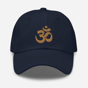 Puede incluir: Una gorra de béisbol azul marino con un símbolo Om bordado en oro.