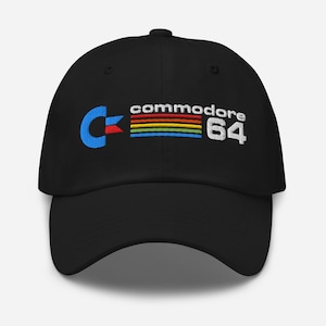 Può includere: Cappellino da baseball nero con il logo Commodore 64 ricamato. Il logo presenta un simbolo 'C' blu, una striscia arcobaleno e la scritta 'Commodore 64' in bianco. Il cappellino ha una visiera curva ed è realizzato in tessuto morbido.