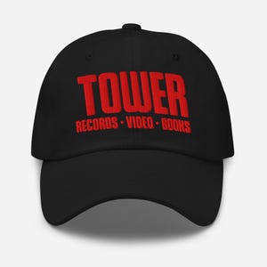 May include: Black baseball cap with the word "TOWER" embroidered in red block letters. Below the word "TOWER" are the words "RECORDS • VIDEO • BOOKS" in red. The cap has a curved brim and an adjustable strap.
