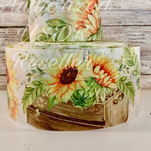 May include: Two rolls of decorative ribbon featuring a watercolor design of sunflowers and greenery. The ribbon has a white background with the words "Authorized USDR Reseller" printed on it. The sunflowers are yellow and orange with brown centers.