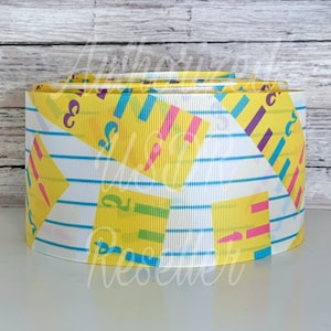 May include: A roll of white and yellow ribbon with a repeating pattern of blue lines and yellow squares with numbers and colorful lines. The ribbon is perfect for crafting and decorating.