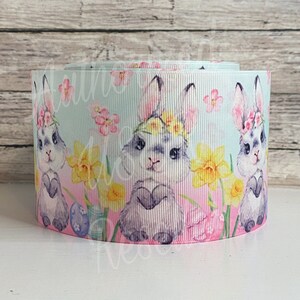 May include: A roll of ribbon featuring an Easter theme. The ribbon has a light blue background with illustrations of bunnies wearing flower crowns, daffodils, and Easter eggs. The ribbon is approximately 2 inches wide.