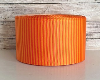 May include: A spool of grosgrain ribbon featuring a repeating vertical striped pattern in vibrant shades of orange and yellow. The ribbon is tightly wound into a cylindrical shape, displaying a consistent ribbed texture across the entire surface.