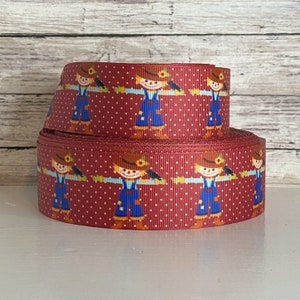 May include: A roll of burgundy ribbon with a repeating pattern of cartoon scarecrows in blue overalls, brown hats, and sunflowers. The ribbon has a white polka dot design.