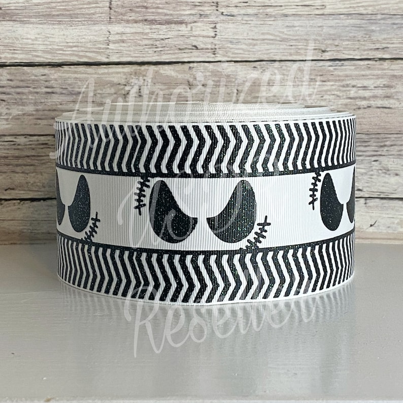 May include: A roll of white ribbon with a black and white Halloween-themed design. The ribbon features a repeating pattern of black zigzags and stylized black eyes with stitched details. The design is suitable for crafts and decorations.
