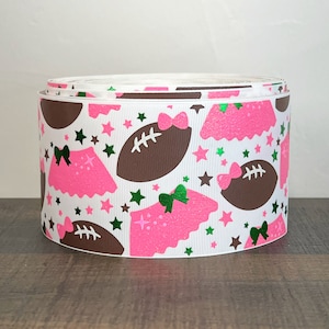 May include: A roll of white ribbon with a repeating pattern of brown footballs, pink glittery skirts, green bows, and stars. The ribbon is suitable for crafting and gift wrapping.