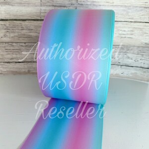 May include: A roll of ombre ribbon with blue and pink stripes. The ribbon is on a white wooden surface.