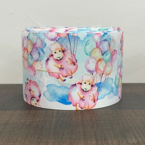 May include: A white ribbon with a repeating pattern of pink sheep holding onto colorful balloons. The sheep are watercolor-style illustrations with pink, blue, and purple hues. The balloons are various shades of pink, blue, purple, and green.