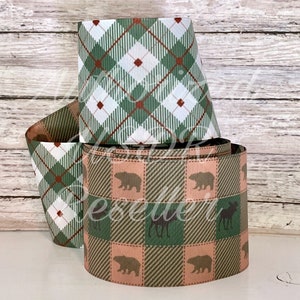 May include: A stack of decorative ribbons with a plaid pattern in green, white, and red. Another ribbon features a repeating pattern of bears and moose in a plaid design with pink and green squares. The ribbons are on a white surface.