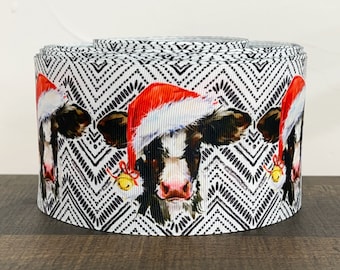 May include: A roll of decorative ribbon featuring a repeating pattern of cow faces wearing Santa hats. The cows are black and white with pink noses, set against a black and white chevron background. Gold bells hang from the hats.
