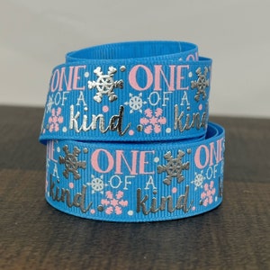 May include: A roll of blue ribbon with the words "ONE OF A KIND" printed in silver and pink. The ribbon features snowflake and dot designs. The ribbon is likely used for crafts, gift wrapping, or decorations.