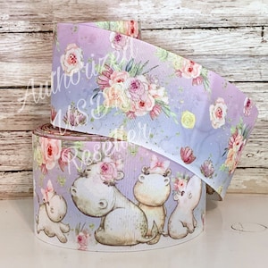 May include: A roll of purple and white ribbon with a floral and hippopotamus print. The ribbon features a watercolor-style design with pink, yellow, and green flowers. The hippos are wearing floral crowns.