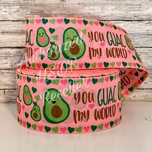 May include: A roll of pink ribbon with a repeating pattern of green avocados, pink and green hearts, and the phrase "You Guac My World" in brown and green. The ribbon is suitable for crafts and gift wrapping.