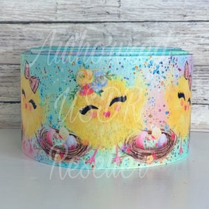May include: A turquoise ribbon with a repeating pattern of yellow chicks holding Easter eggs in nests. The chicks have flowers on their heads and are surrounded by a light blue and pink background with white speckles.
