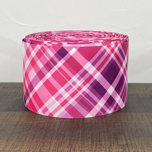 May include: Pink and purple plaid ribbon with a white stripe. The ribbon is rolled up and ready to be used for crafting or decorating.