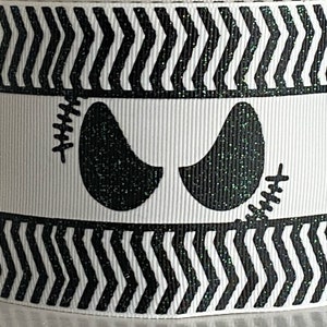 May include: White ribbon with a black and white zigzag pattern and a central band featuring a pair of black glittery eyes with stitched details. The ribbon is likely for crafting or decoration.