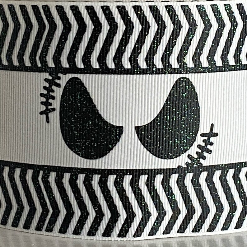 May include: White ribbon with a black and white zigzag pattern and a central band featuring a pair of black glittery eyes with stitched details. The ribbon is likely for crafting or decoration.