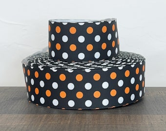 May include: Two rolls of black ribbon with a repeating pattern of white and orange polka dots. The ribbon is stacked on top of each other, with the larger roll at the bottom and the smaller roll on top. The ribbon is on a dark brown surface.
