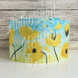 May include: A roll of blue and yellow ribbon with a watercolor floral print. The ribbon features yellow flowers with black centers and a light blue background with gold glitter.