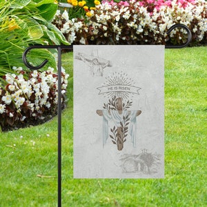 May include: A garden flag with a white background and a brown wooden cross with a white cloth draped over it. The cross is surrounded by green leaves and the text "He is Risen" is written in black lettering.