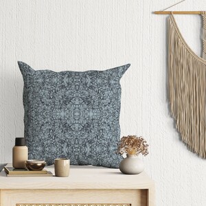 May include: A blue patterned throw pillow with a geometric design sits on a wooden table with a woven rattan front. A small vase with dried flowers and a bottle are also on the table. A macrame wall hanging is in the background.