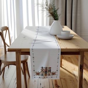 May include: White table runner with a festive design featuring three nutcracker figures in red, blue, and green. The runner has a white background with a black and red geometric border.