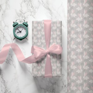 May include: A gift box wrapped in gray wrapping paper with a pink ribbon and a pattern of white bunnies. The box is sitting on a white marble surface next to a green alarm clock.