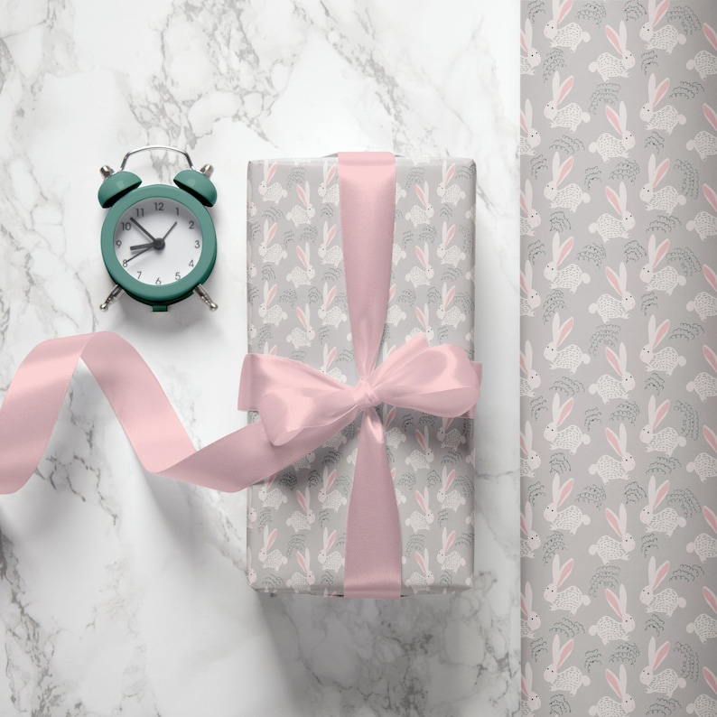 May include: A gift box wrapped in gray wrapping paper with a pink ribbon and a pattern of white bunnies. The box is sitting on a white marble surface next to a green alarm clock.