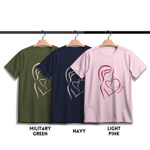 May include: Three t-shirts with a graphic of a mother and child in a heart shape. The t-shirts are in military green, navy blue, and light pink. The pink t-shirt has the text "#momlife" in burgundy.