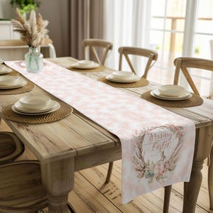 May include: A white table runner with a pink floral pattern and the text "Happy Easter" in a wreath. The runner is on a wooden table with two white bowls and a brown placemat.