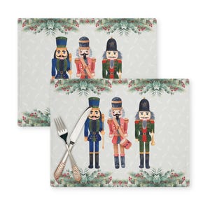 Op de afbeelding: Set van twee kerst-placemats met een aquarel-illustratie van notenkrakersoldaten in blauw, rood en groen op een witte achtergrond met een groene en witte bloemenrand.