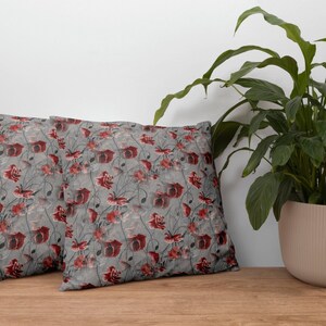 May include: Two square throw pillows with a floral pattern. The design features red flowers and dark stems on a gray background. The pillows are placed on a wooden surface, with a potted plant in the background.