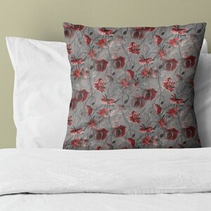 May include: A decorative throw pillow featuring a floral pattern of red poppies on a gray background. The pillow is square and rests on a white pillow and bed linens. The design includes stems and leaves.