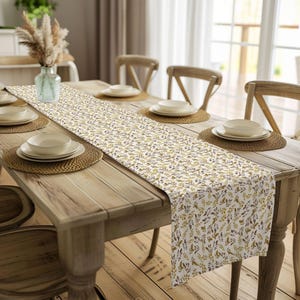 May include: A white table runner with a brown and purple floral pattern. The runner is laid on a wooden table with two place settings.
