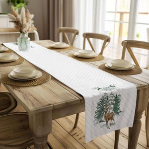 May include: White table runner with a green and brown deer and forest scene. The runner has a white background with thin grey stripes. The runner is on a wooden table with place settings.