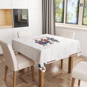 May include: White tablecloth with a festive Christmas design featuring a group of nutcracker figures and the text "Happy Holidays". The tablecloth is laid on a wooden table with two chairs.