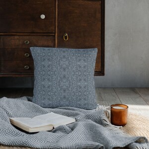 May include: A gray throw blanket with a black and white diamond pattern is draped over a dark wooden cabinet. A gray patterned pillow sits on the blanket. A candle sits on a woven rug.