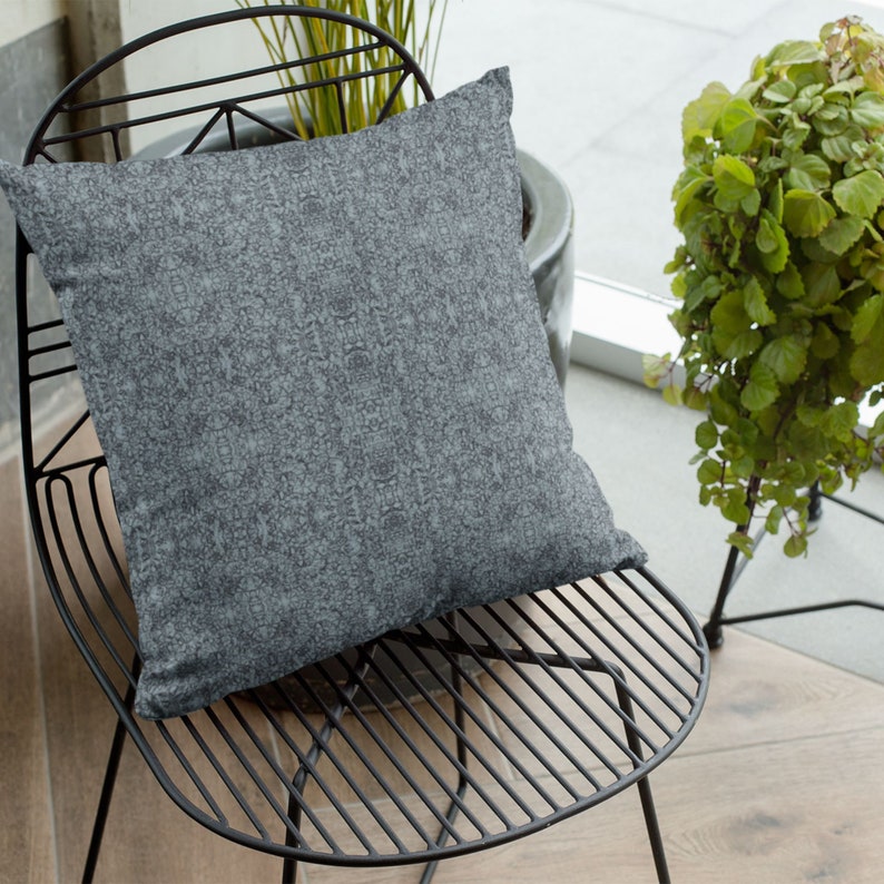 May include: A gray and white patterned throw pillow on a black metal chair. The chair has a slatted design and is in a room with a window and plants.