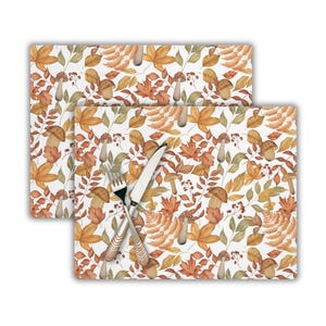 May include: Two brown and orange watercolour placemats with a pattern of leaves, mushrooms, and berries. The placemats are set on a white background with a fork and knife on top.
