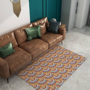 以下が含まれることがあります： 黄、赤、青の色合いの模様のある敷物の上に、緑と茶色のクッションを置いた茶色の革のソファが置かれています。敷物は、円とアーチが繰り返される幾何学的な模様をしています。