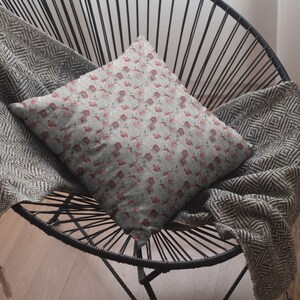 May include: Square throw pillow with a floral pattern in shades of red and gray. The pillow is resting on a black chair with a woven design, and a gray blanket is draped over the chair.