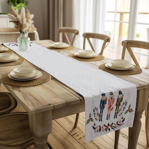 May include: White table runner with a white background and a repeating pattern of small white dots. The runner features a colourful illustration of three nutcracker figurines with holly and berries.