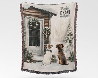Puede incluir: Una manta tejida con una escena invernal que muestra la puerta de una casa, dos coronas, un pino nevado y dos perros sentados en la nieve. El texto "Baby It's Cold Outside" está impreso en letra cursiva negra. La manta tiene bordes con flecos.